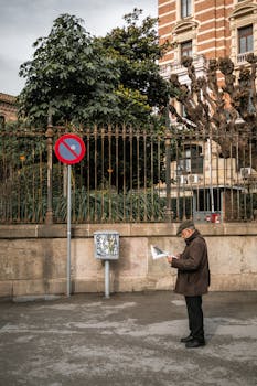 An elderly man reads a newspaper while standing on a Barcelona street, next to a 'no parking' sign.