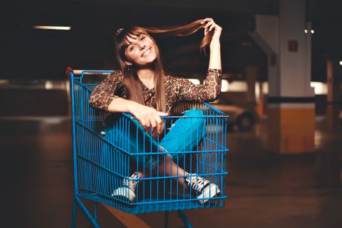 Free Smiling woman in leopard print top and jeans sitting playfully in a blue shopping cart. Stock Photo
