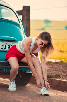 Young woman ties shoes next to a classic blue car on a sunny rural road.