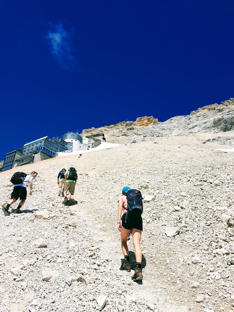Group Of Tourists With Backpacks Climbing Mountain Under Blue Sky