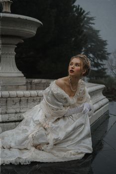 A woman in a vintage white gown poses thoughtfully by a stone fountain outdoors.