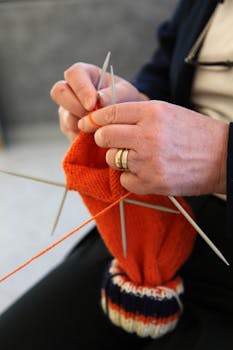 A detailed view of hands knitting with orange yarn using multiple needles.