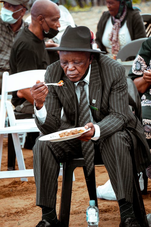 Man in Black Suit Jacket and Black Pants Sitting on White Plastic Chair while Eating Meal