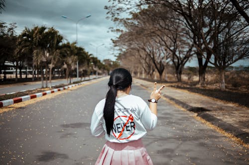 Woman Wearing White Long-sleeved Shirt and Pink Pleated Skirt Standing on Asphalt Road Between Green Trees Under Gray Sky at Daytime