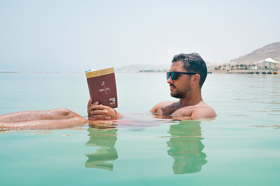 Free Man Wearing Sunglasses Reading Book on Body of Water Stock Photo