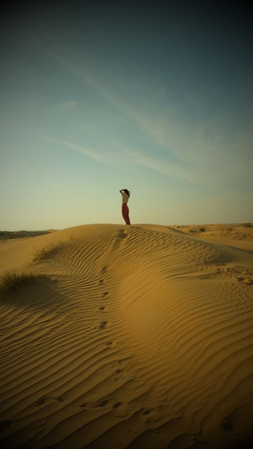 Free A woman stands on sand dunes with a clear blue sky, capturing the serene desert dawn. Stock Photo