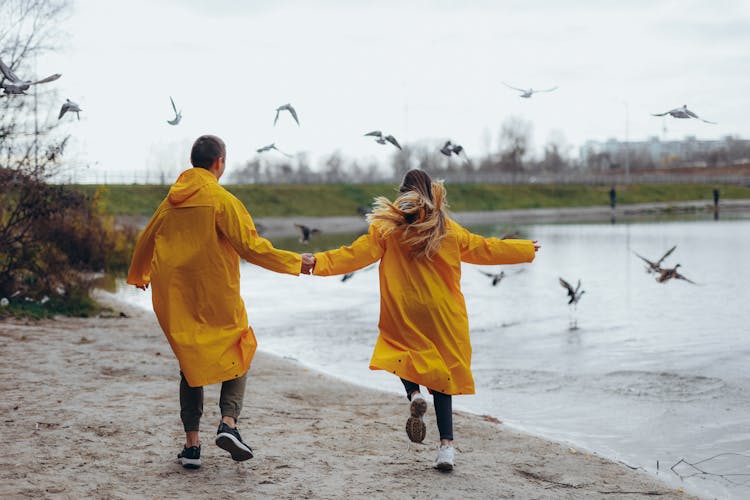 Couple In Yellow Raincoats Running On The Beach And Holding Hands