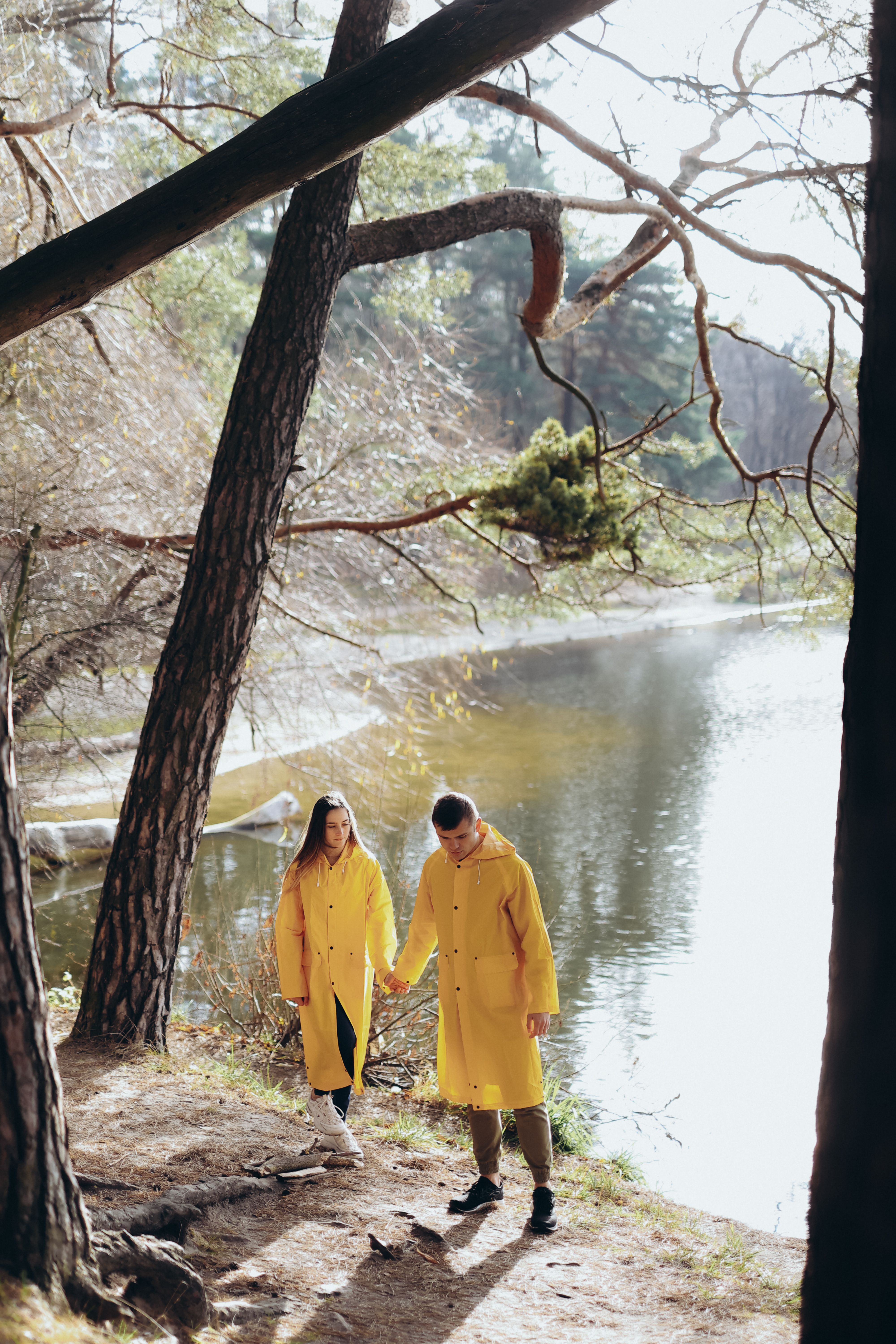 A couple holding hands, walking along a lakeside in bright yellow raincoats, surrounded by trees.