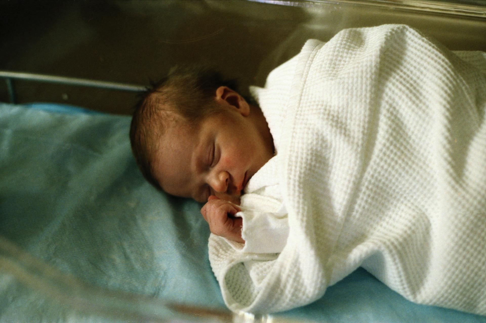 A peaceful newborn baby sleeping wrapped in a white blanket in a hospital setting.