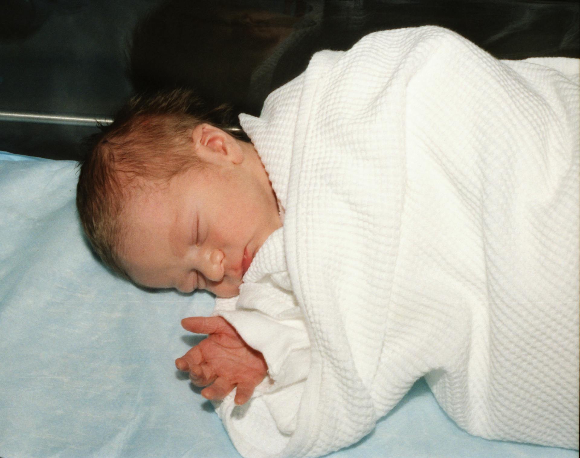 A serene newborn baby peacefully sleeping, wrapped in a white hospital blanket on a light blue sheet.