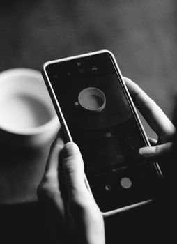 Monochrome close-up of hands holding smartphone capturing a coffee cup.