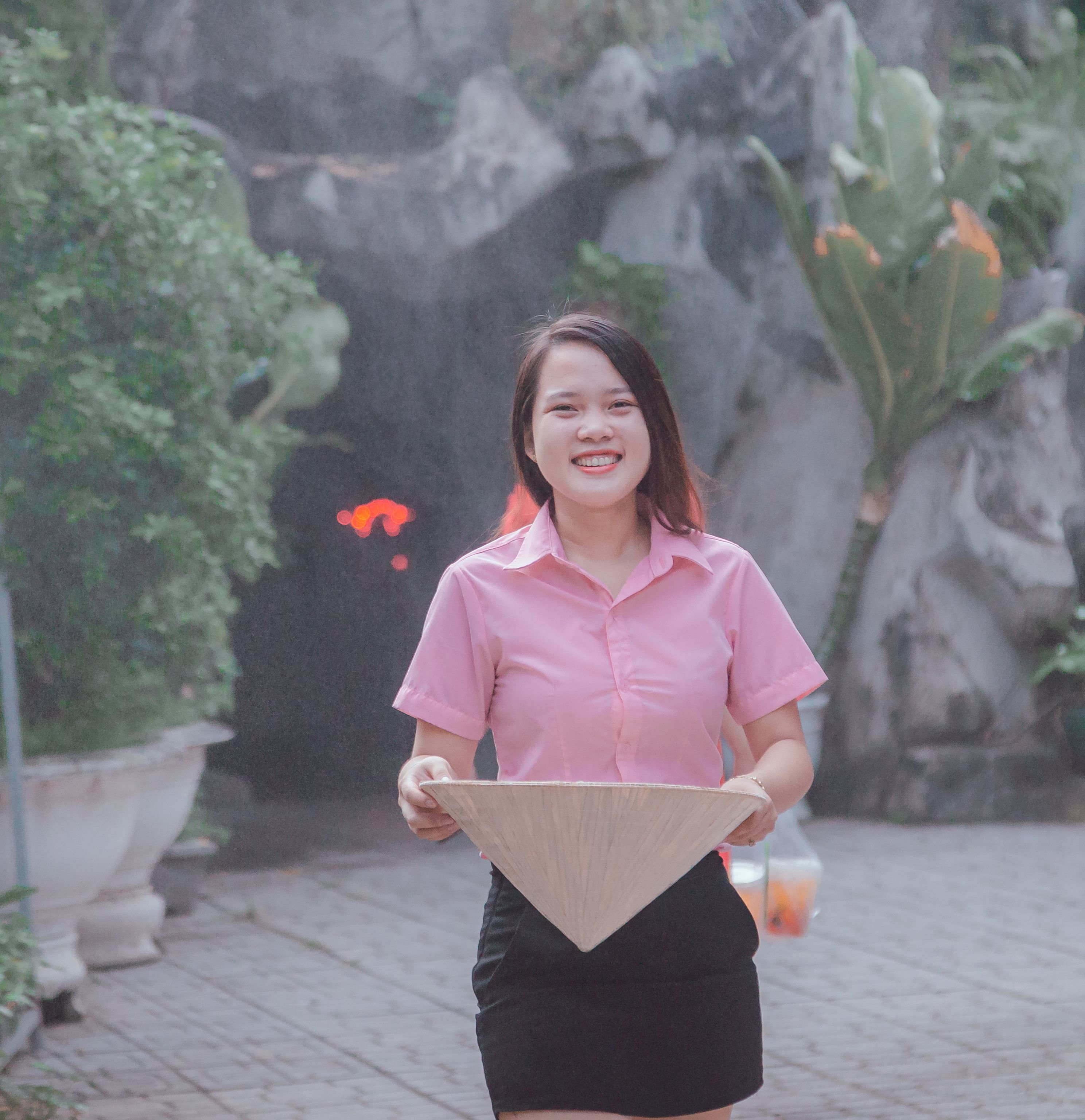 Asian woman smiling outdoors in a pink shirt holding a conical hat, captured in a serene garden setting.