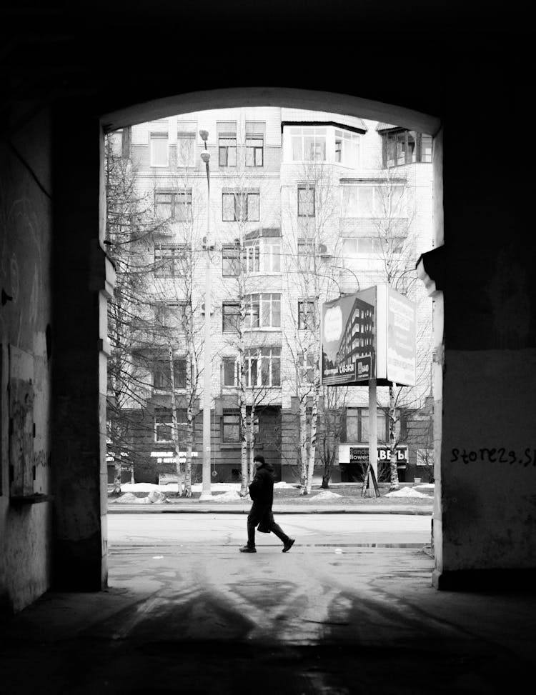 Grayscale Photo Of A Person Walking On Sidewalk Near Building