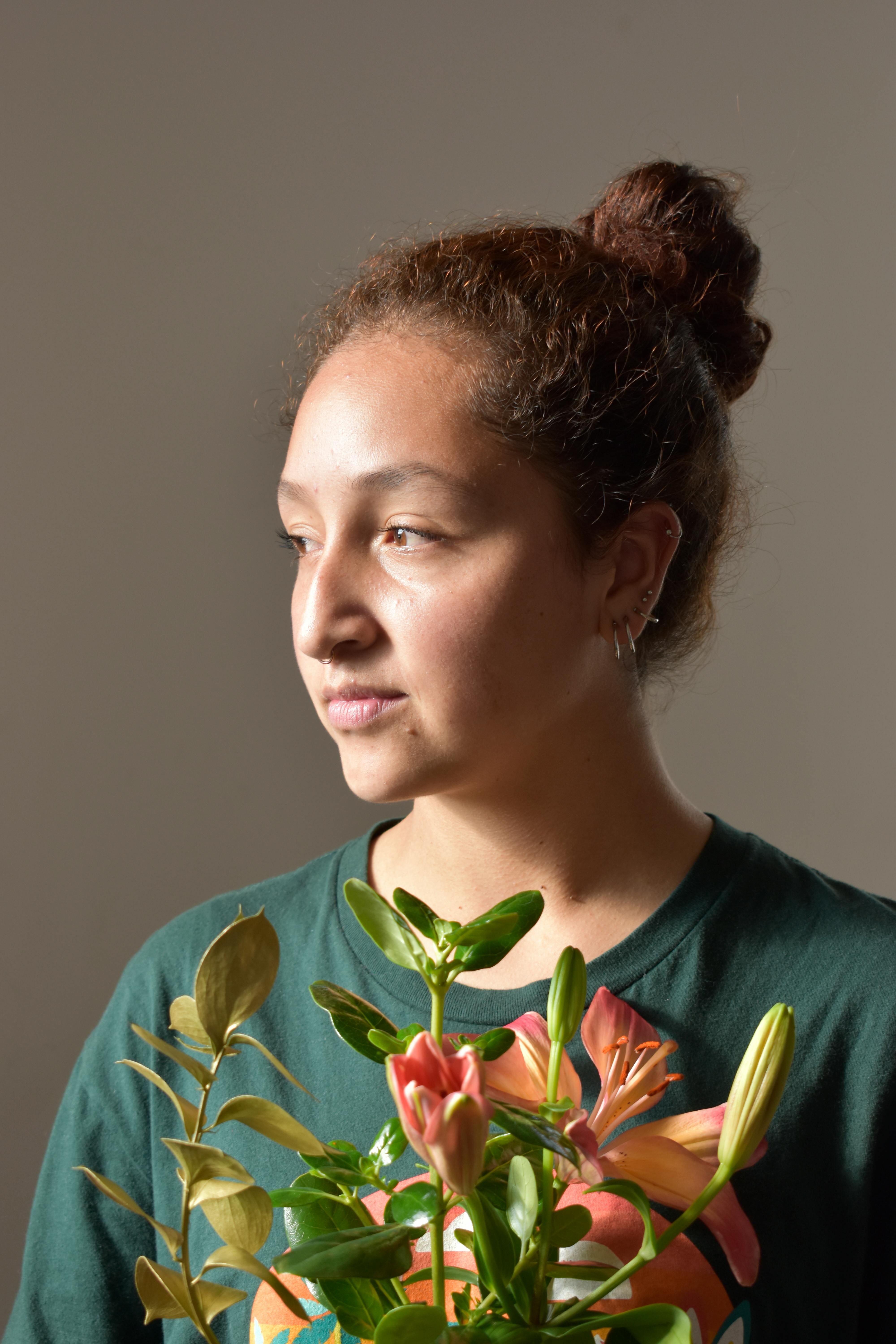 A young woman in a green shirt holding pink lilies, gazing thoughtfully indoors.