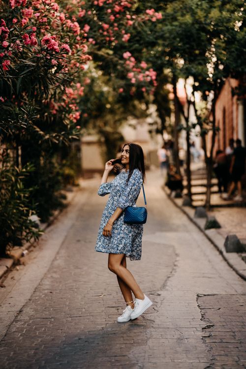 Free Elegant woman in a floral dress walks down a charming street lined with vibrant flowers. Stock Photo