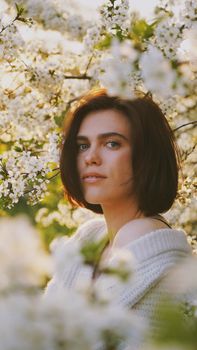 A woman in a white sweater stands among blooming trees, capturing the essence of spring.