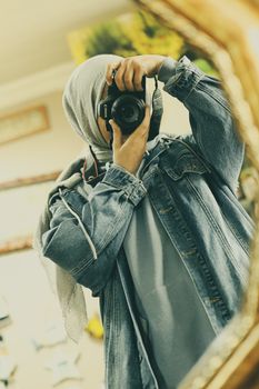 A woman in a hijab and denim jacket takes a selfie with a DSLR camera. Indoors shot.