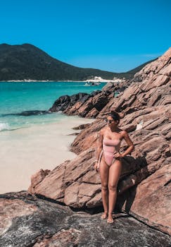Woman in swimsuit standing on rocky beach with turquoise water and clear sky.