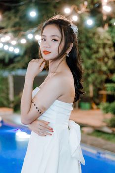A young woman in a white dress poses elegantly by a lit-up poolside, exuding charm and grace.