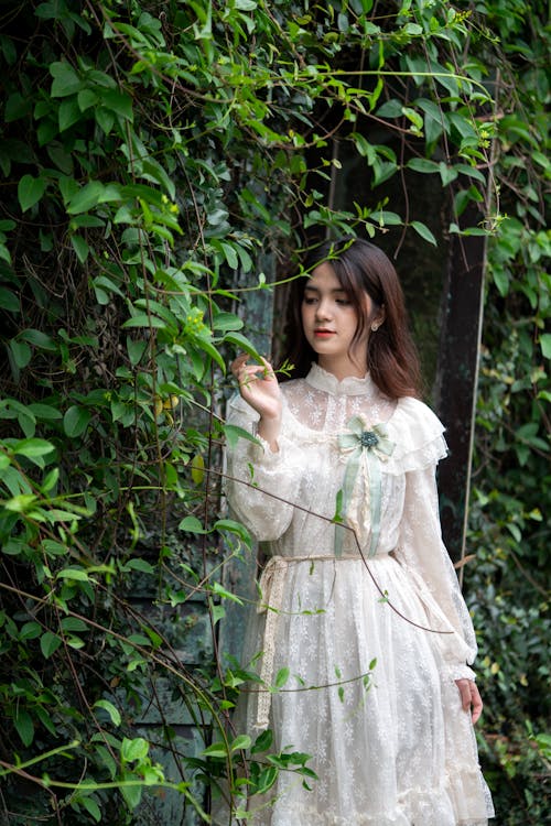 Woman in White Lace Dress Standing Beside Green Plants
