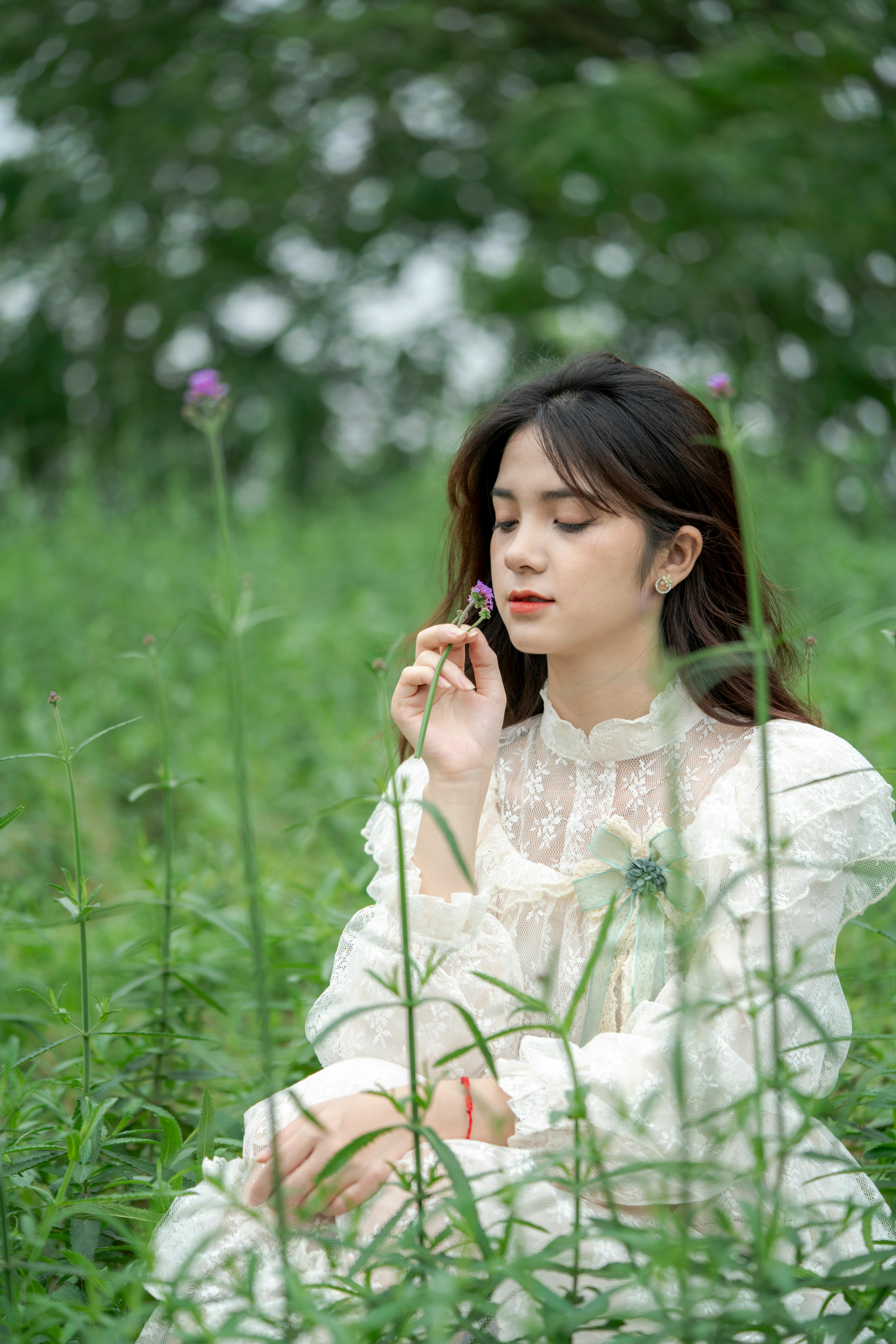 A woman in a white lace dress peacefully sitting in a lush green field with eyes closed, holding a flower.