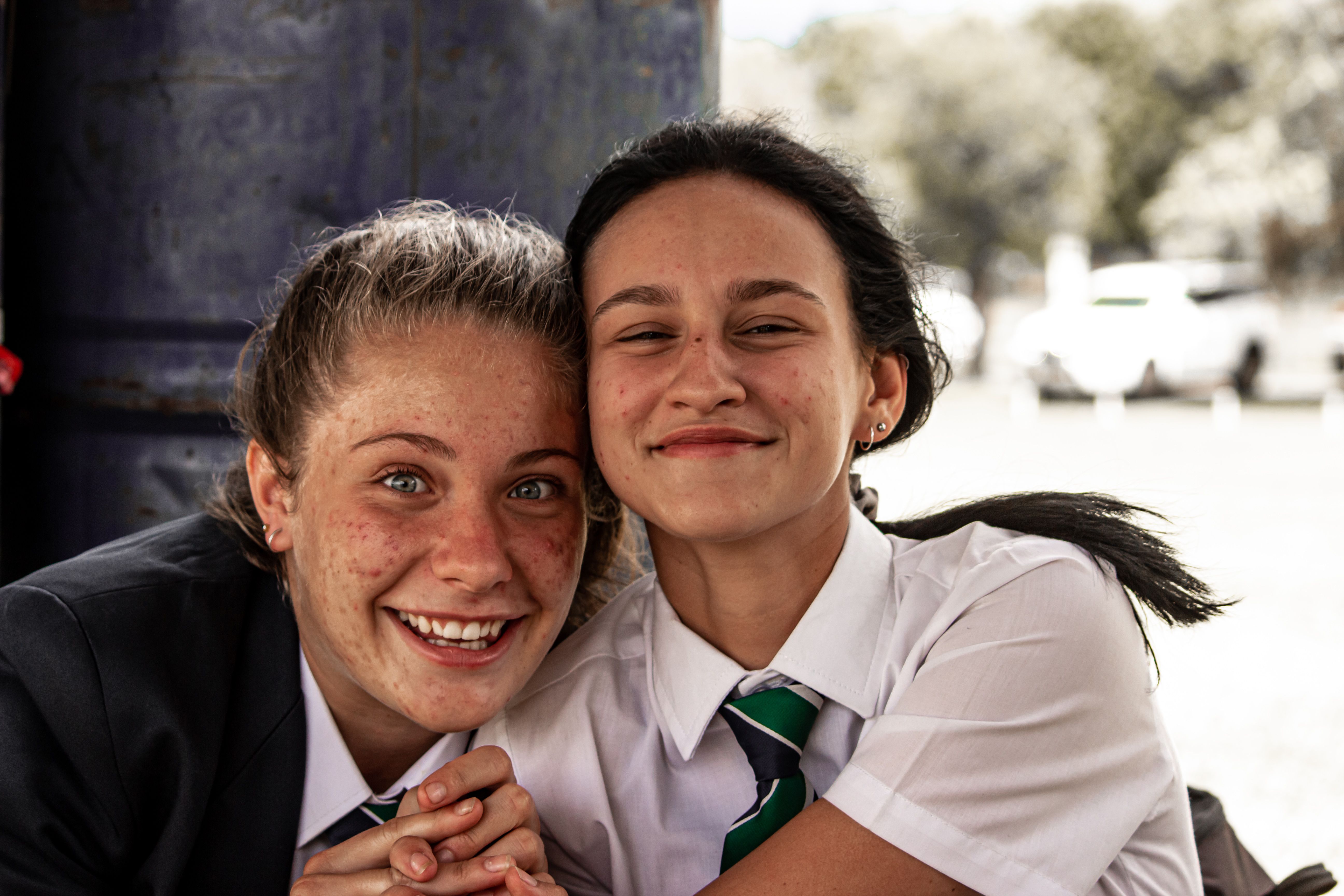 Two smiling teenage girls in school uniforms standing outdoors together, laughing and happy