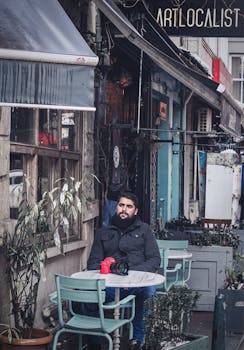 A bearded man in a black jacket with a camera sits at an outdoor cafe surrounded by plants.