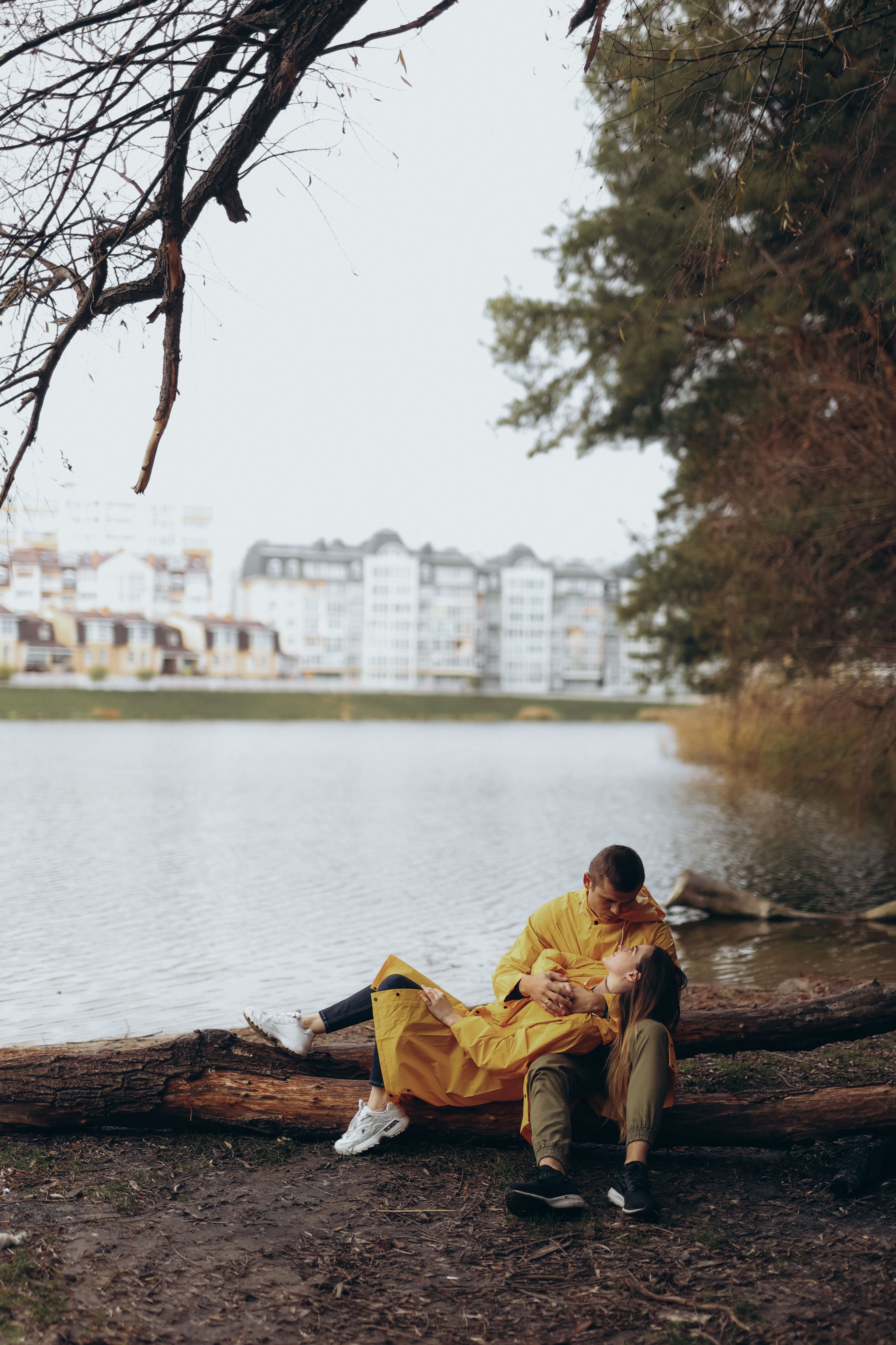 Couple in yellow raincoats sitting on a log by the lake, enjoying a romantic moment in nature.