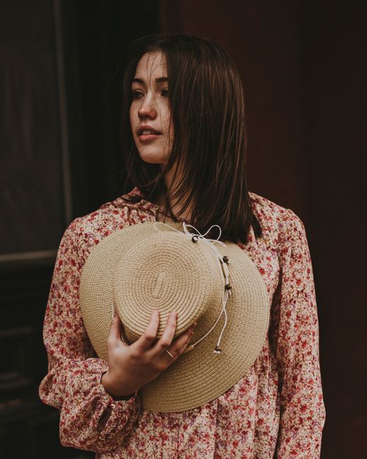 Stylish woman in a floral dress holding a straw hat, exuding a vintage and casual vibe.