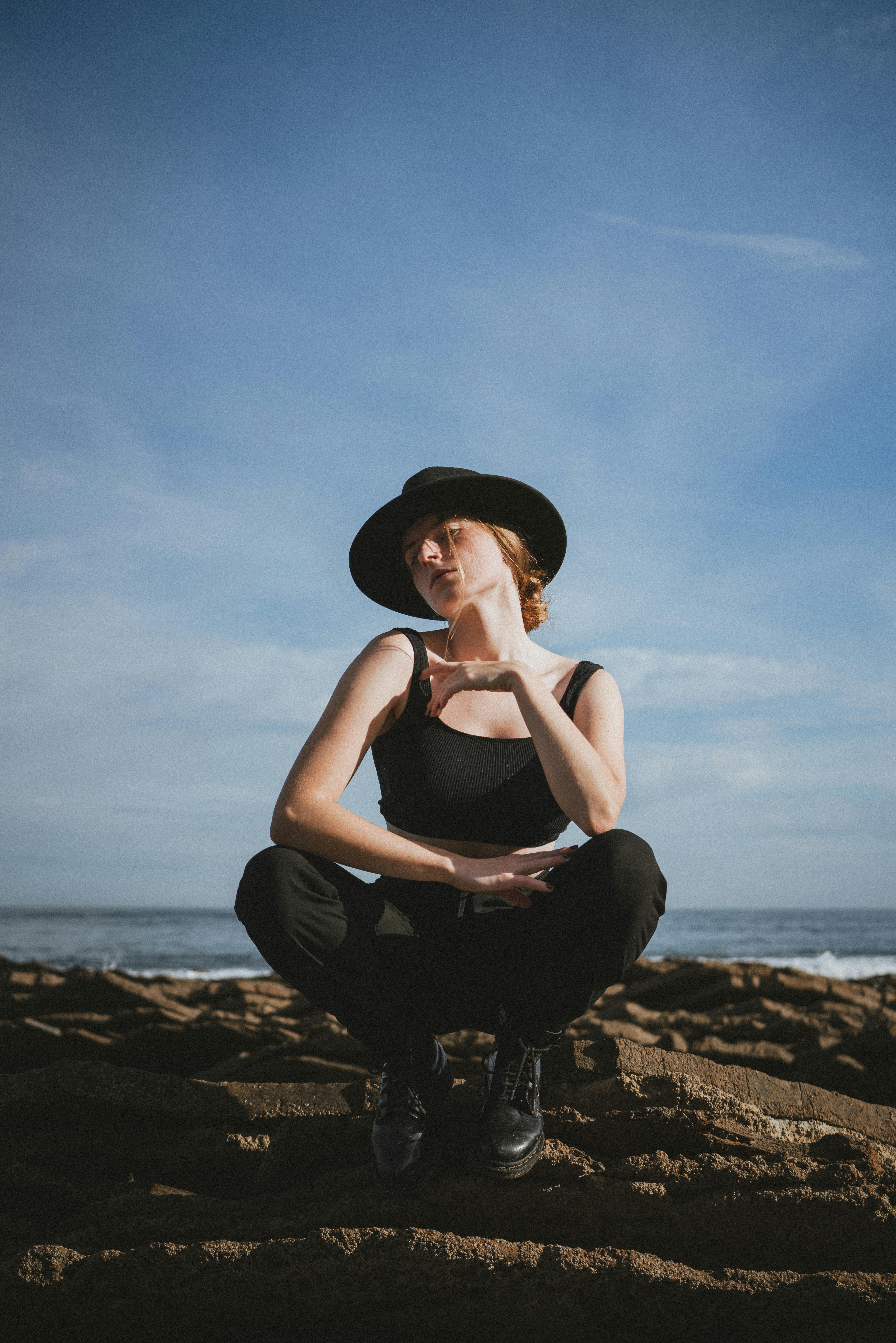 A woman poses in a black hat on a rocky beach with the sea in the background.