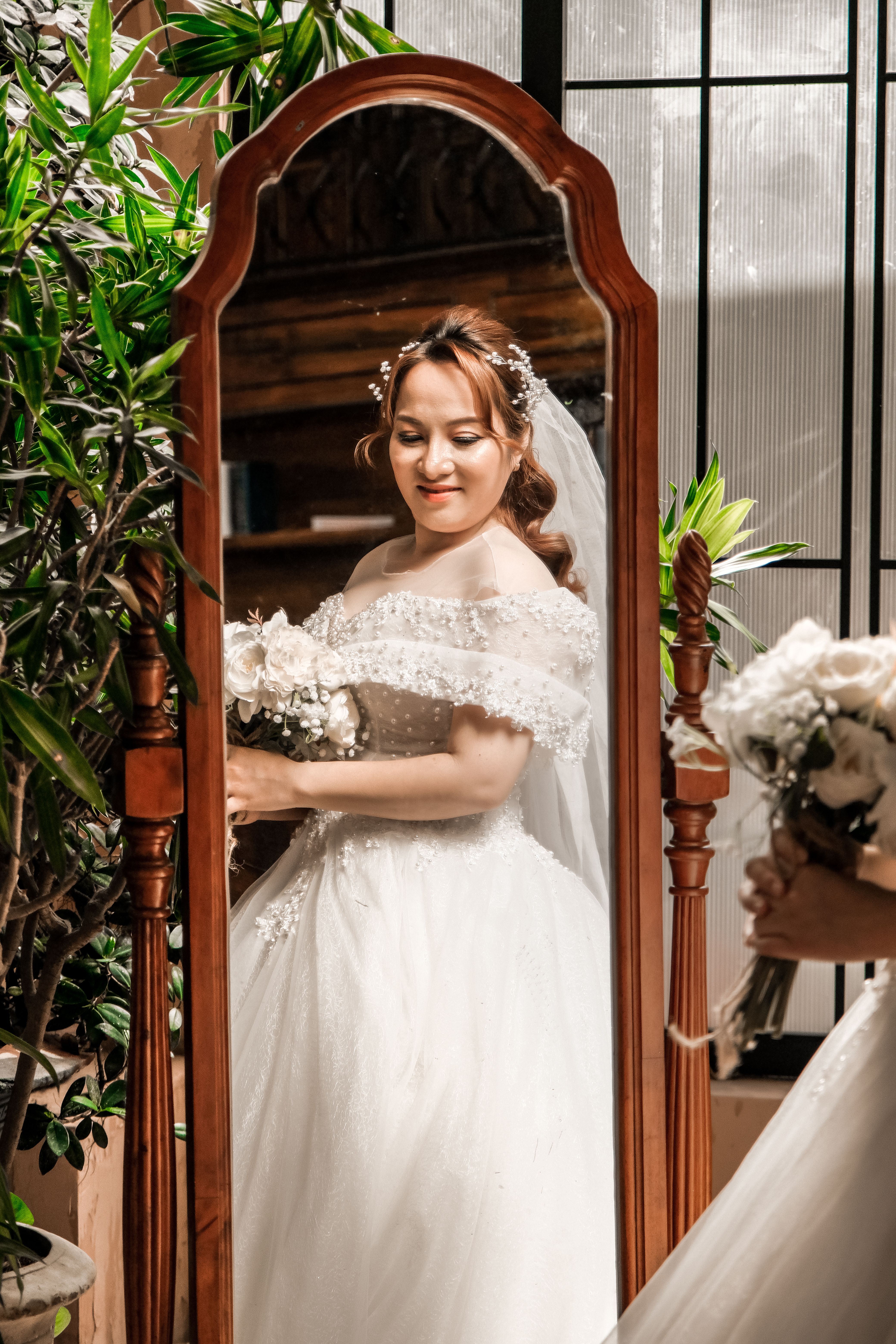 A bride in a wedding dress holding a bouquet looks at her reflection indoors.