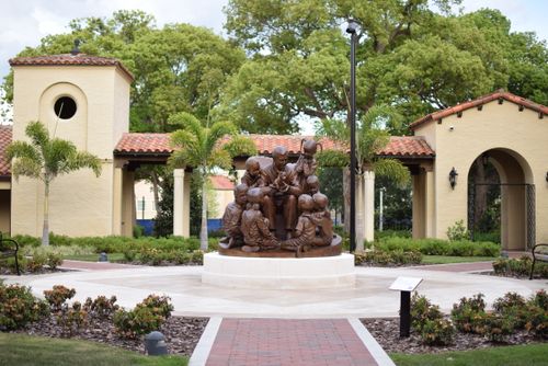 Free Bronze sculpture in a park setting surrounded by lush trees and architecture. Stock Photo