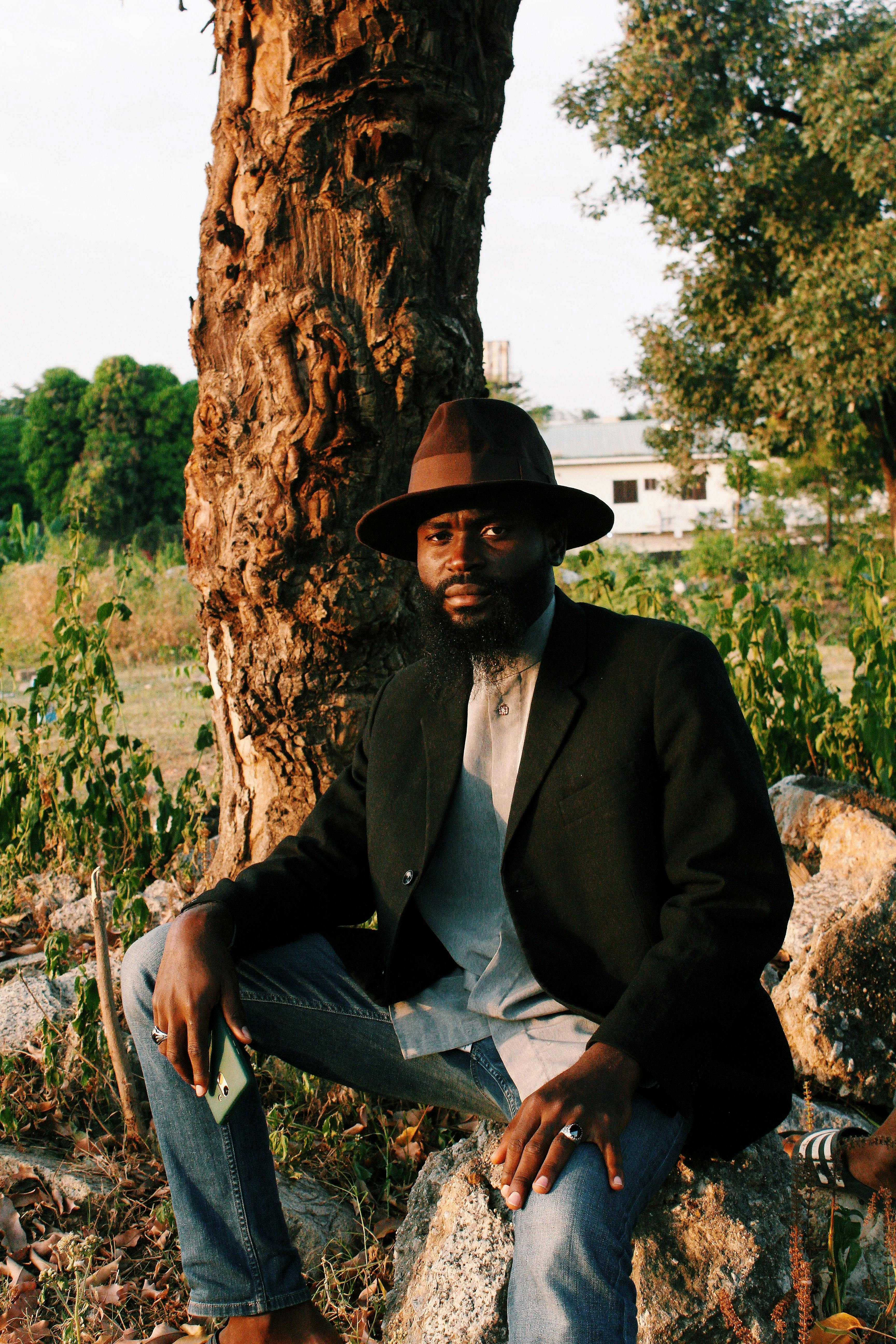 A man in a hat sits near a tree in Abuja, Nigeria, showcasing natural beauty and style.