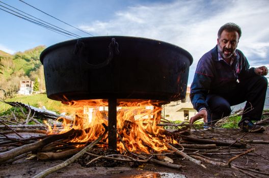 A man tends to an outdoor fire cooking in a large cauldron amidst a natural setting.