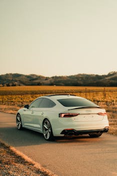A sleek white Audi car on a winding road through picturesque cropland during sunset.