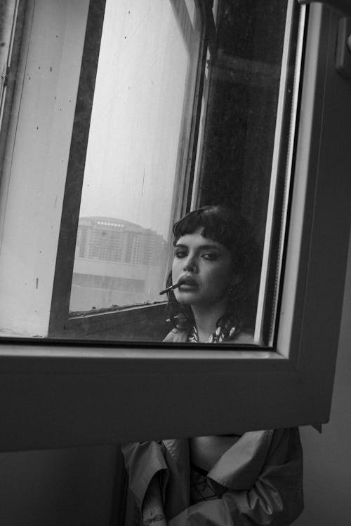 Grayscale Photo of Woman Standing near the Glass Window