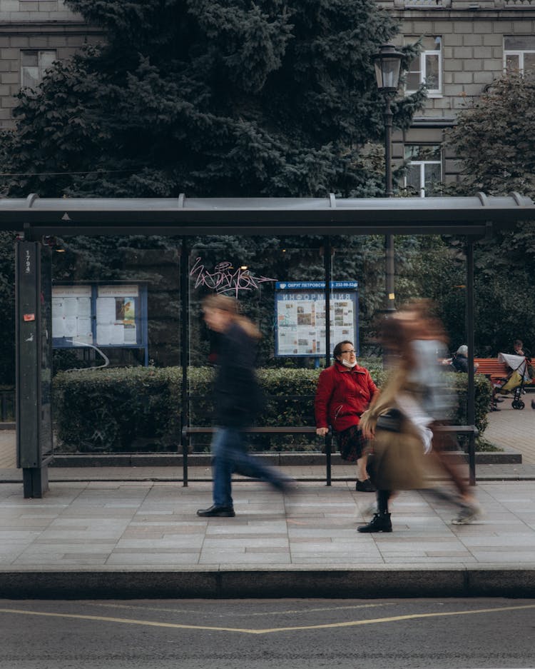 Woman In Red Coat Sitting On Bus Stop With People Passing By