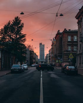 A vibrant urban street scene at sunset featuring traffic, buildings, and a silhouette skyline.