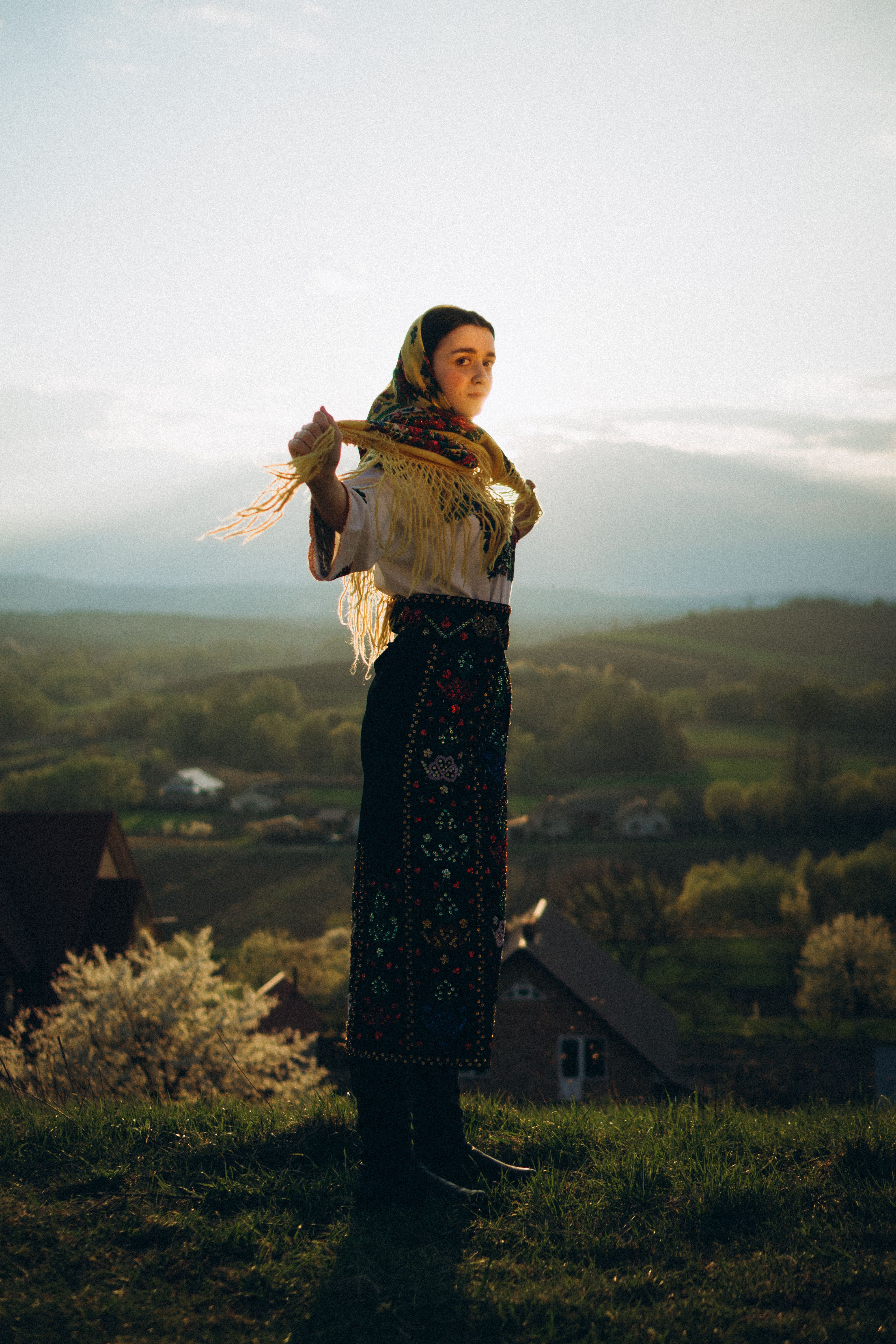 Portrait of a young woman wearing traditional clothing with a scenic countryside backdrop at sunset.