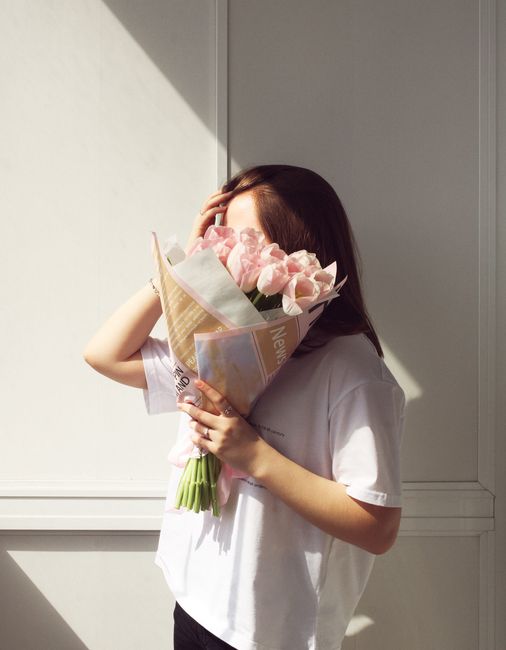 A woman in a white shirt holds a bouquet of pink tulips indoors, illuminated by soft sunlight.