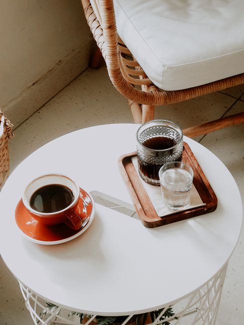 A rustic wicker chair with a coffee setup on a white table in a serene setting.