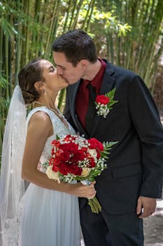 Bride and groom share a kiss outdoors with vibrant red bouquet in hand.