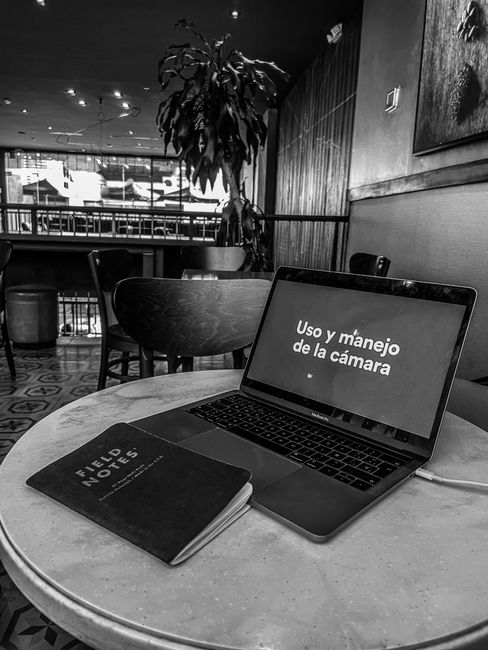 Grayscale image of a laptop displaying 'Uso y manejo de la cámara' on a cafe table with a notepad.