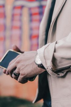 A stylish man in a beige jacket holds a smartphone, showcasing a wristwatch and a ring.