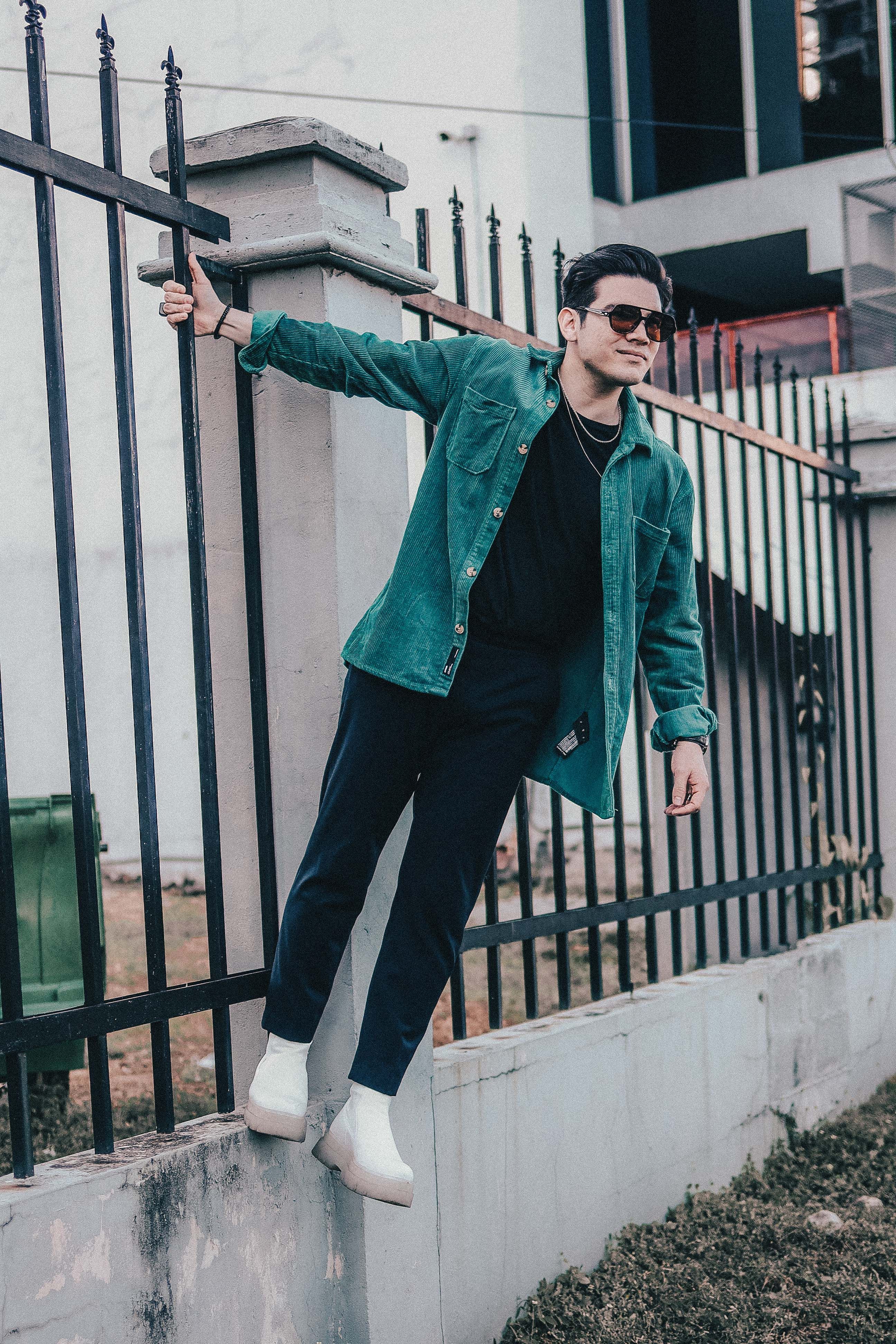 Stylish man in green jacket leaning on metal railings outdoors in urban area.
