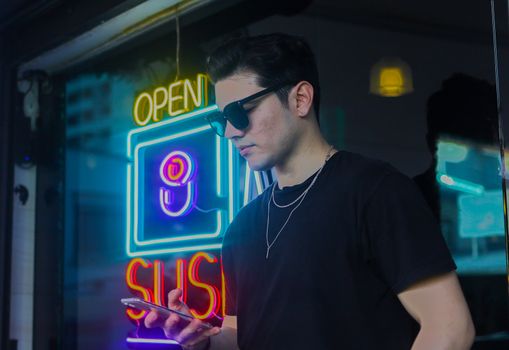 Young man in sunglasses stands near neon sushi sign, creating a modern and stylish urban vibe.