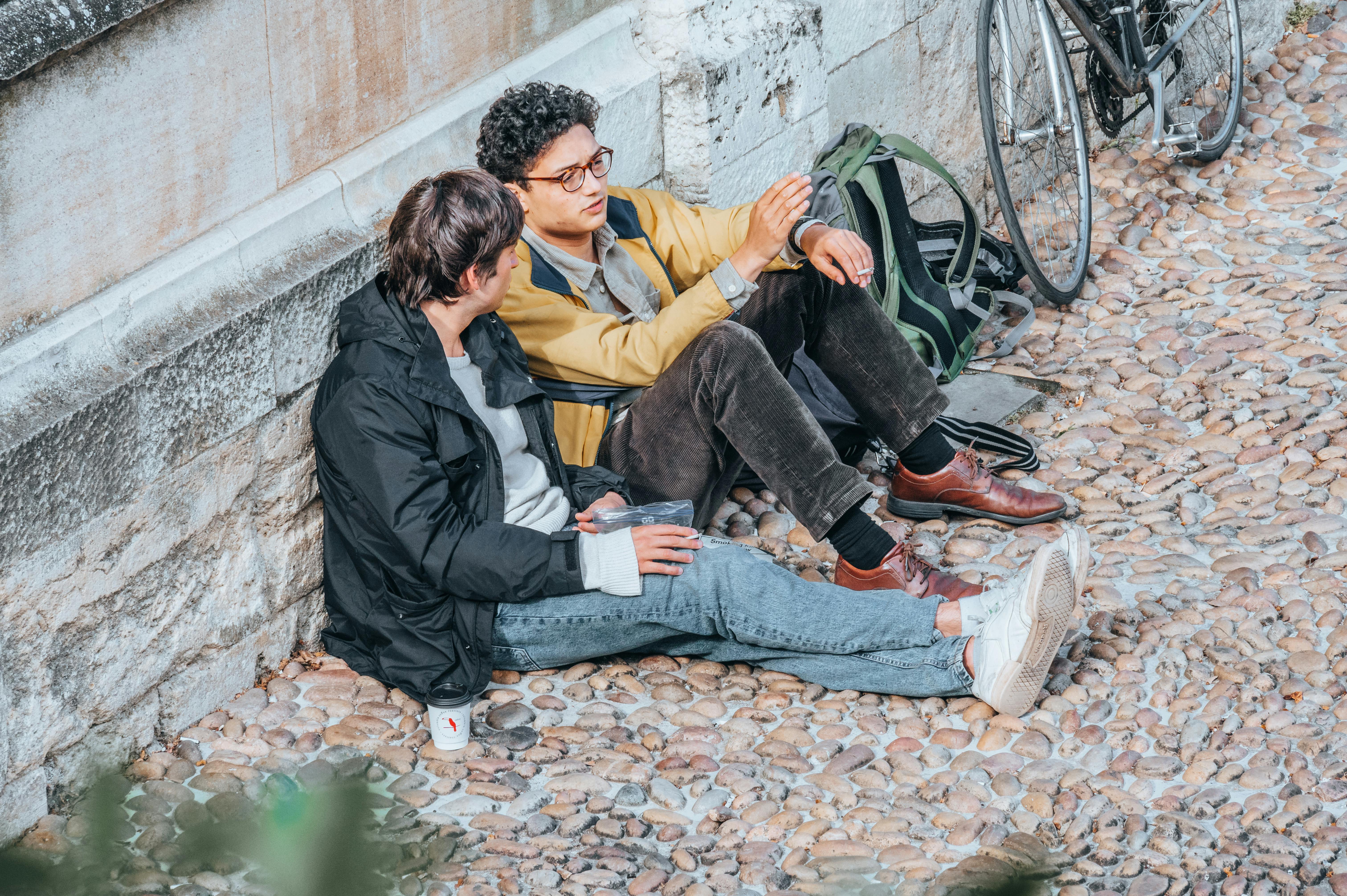 Two young men sit and chat on cobblestones in Oxford, UK, enjoying a casual outdoor conversation.