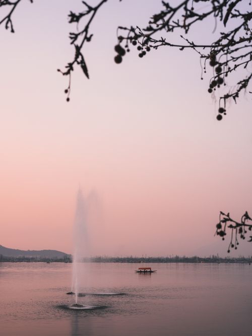 Free A serene view of Dal Lake at sunrise, featuring a lone boat and a fountain amidst a peaceful pink sky. Stock Photo