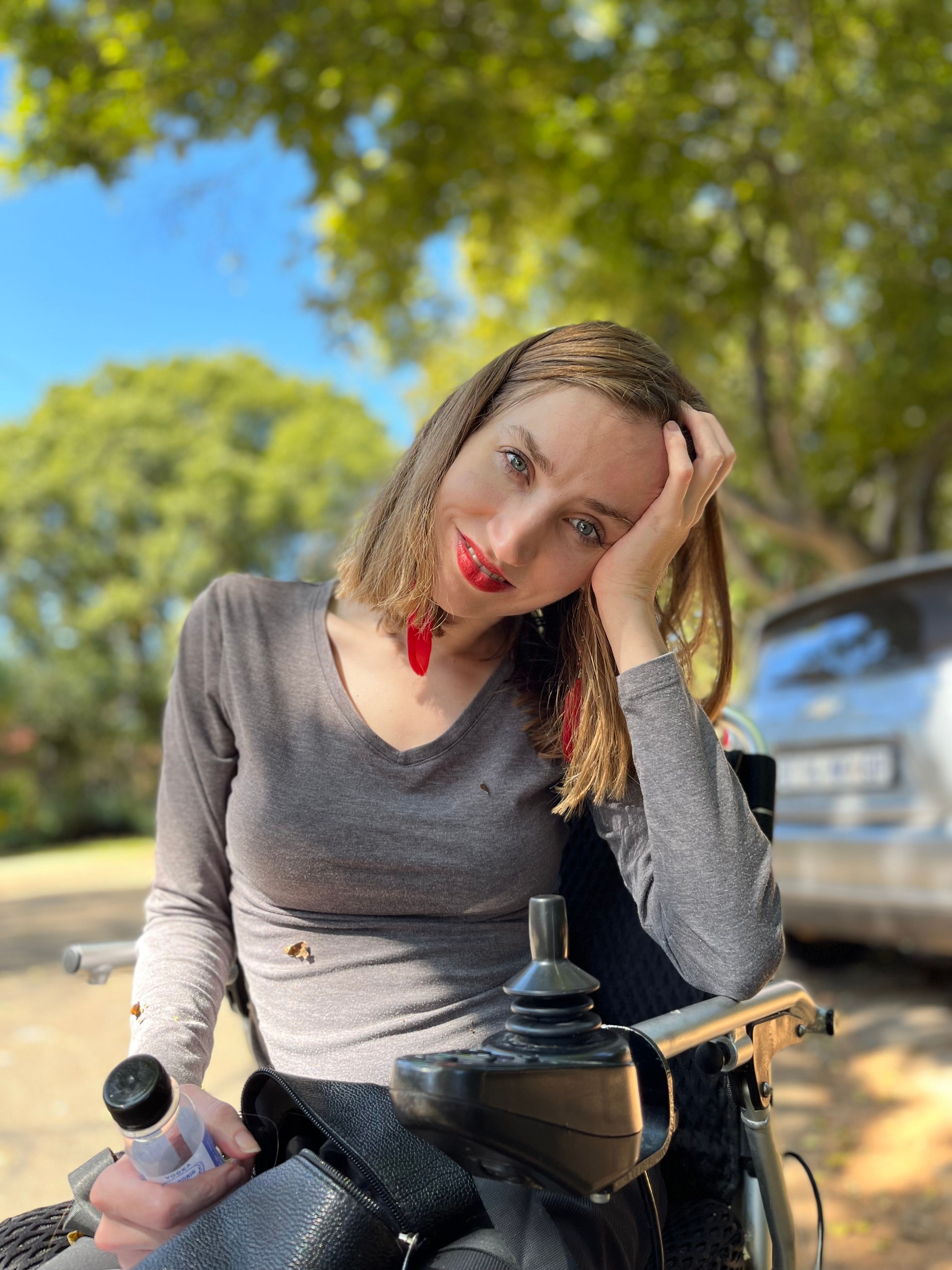 Smiling woman sitting in wheelchair outdoors, enjoying a sunny day under green trees.