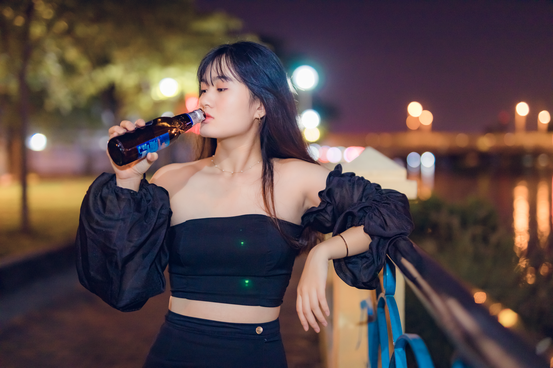 A fashionable woman enjoys a bottled drink by a riverfront at night, showcasing chic style and relaxation.